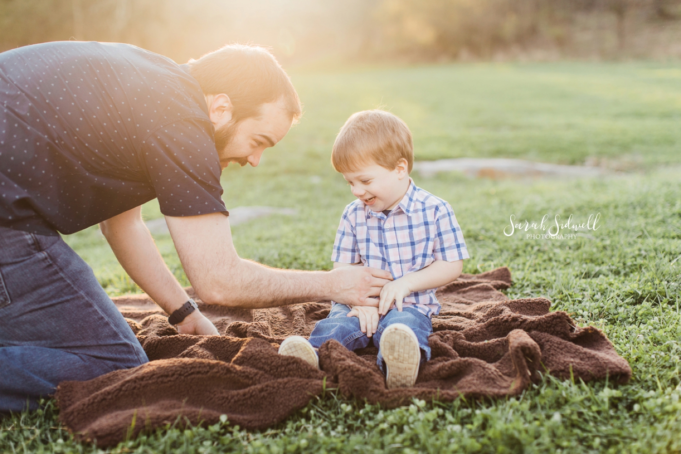 A father plays with his son who sits on a blanket. 