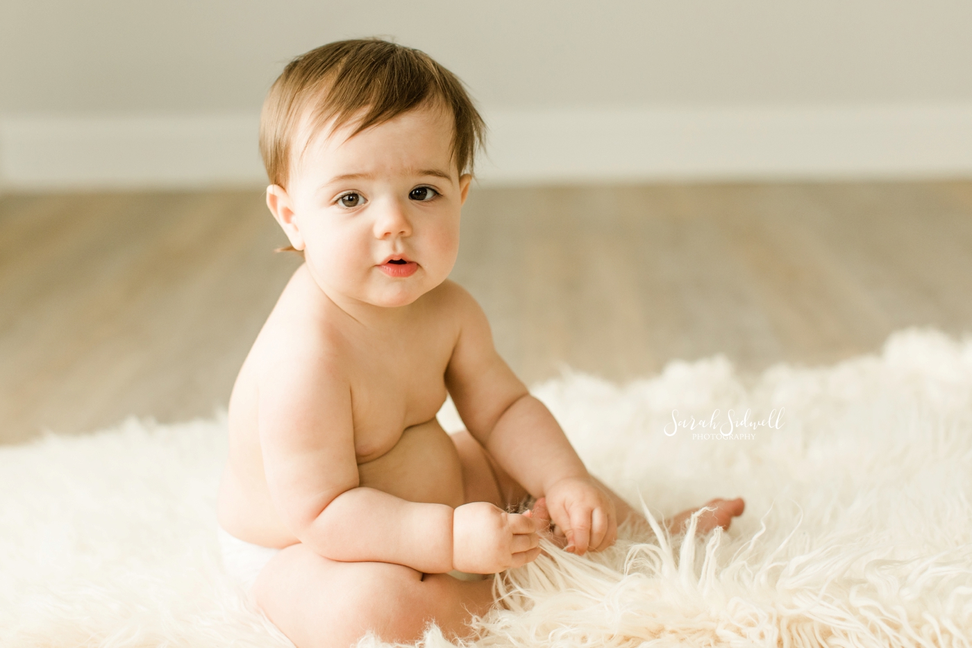 A baby sits on a white rug. 