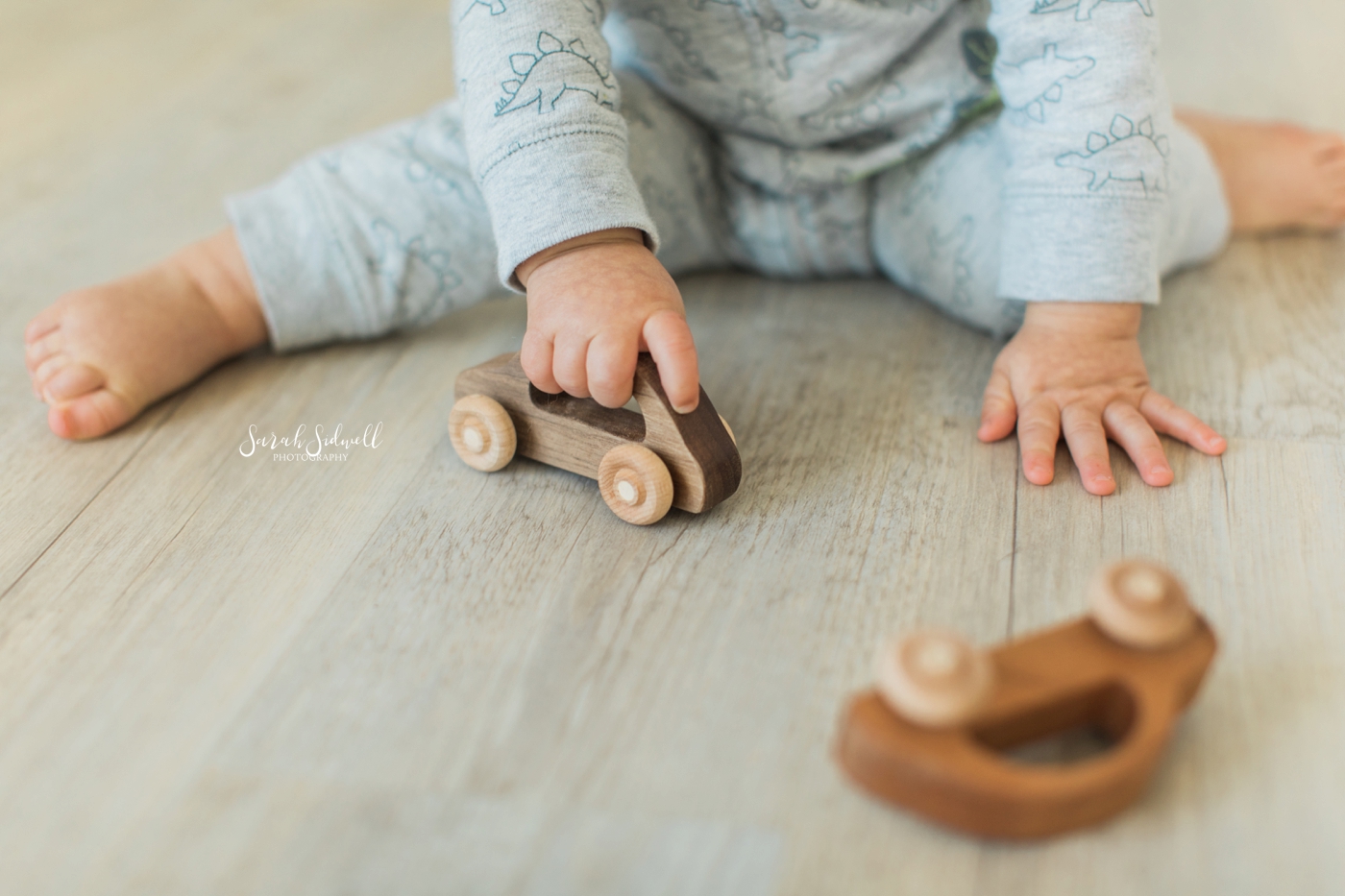 A baby sits on a grey floor and plays with a wooden car. 