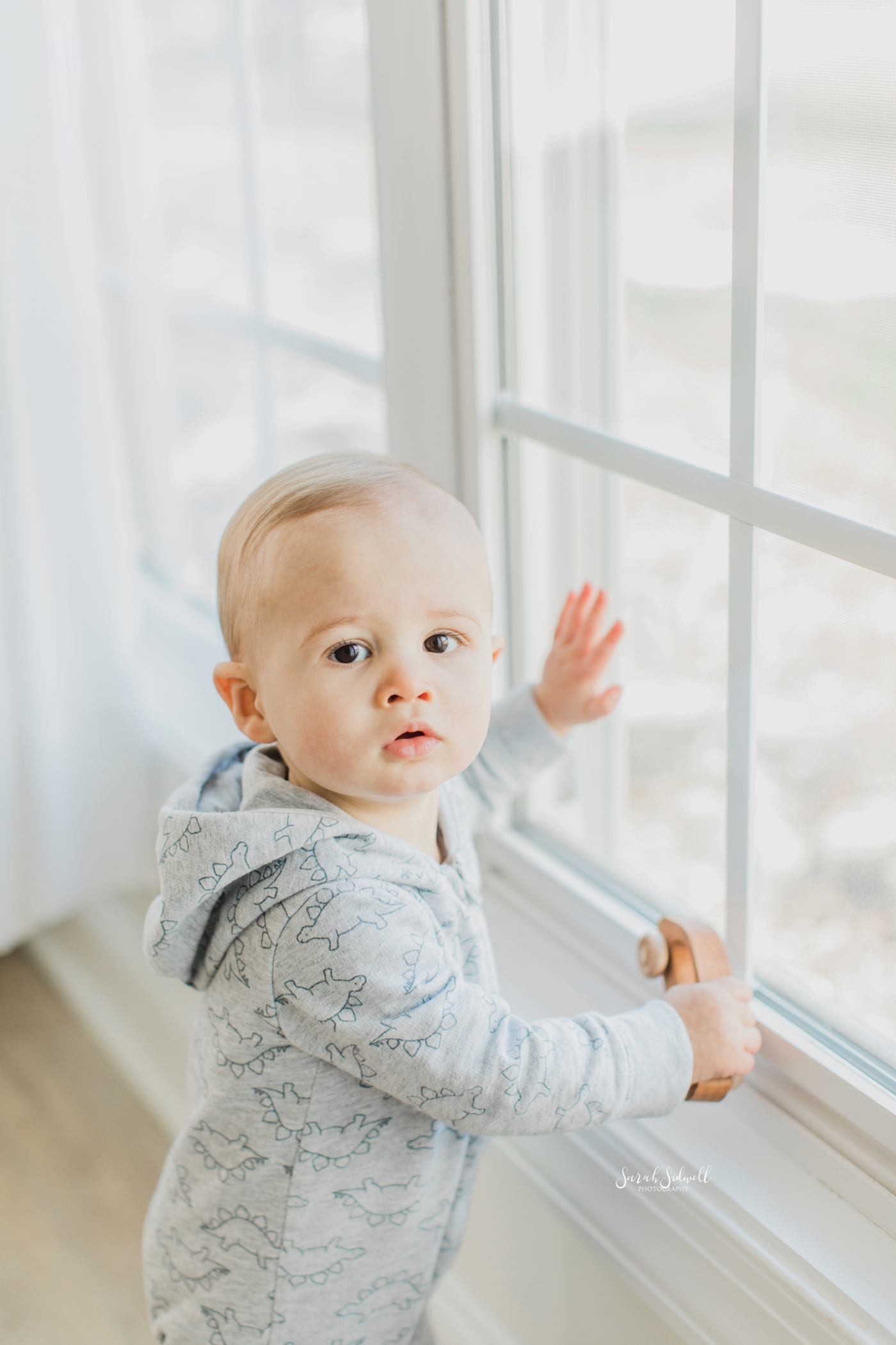 A baby wearing a blue jumper stands at a window and puts his hands up to it. 