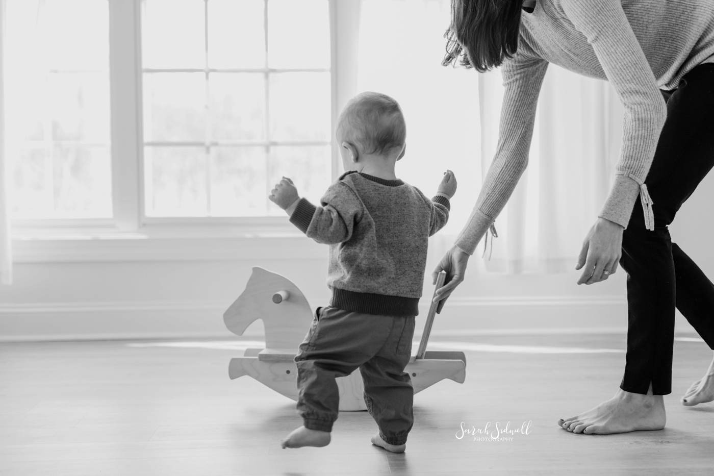 A baby walks towards a wooden rocking horse. 