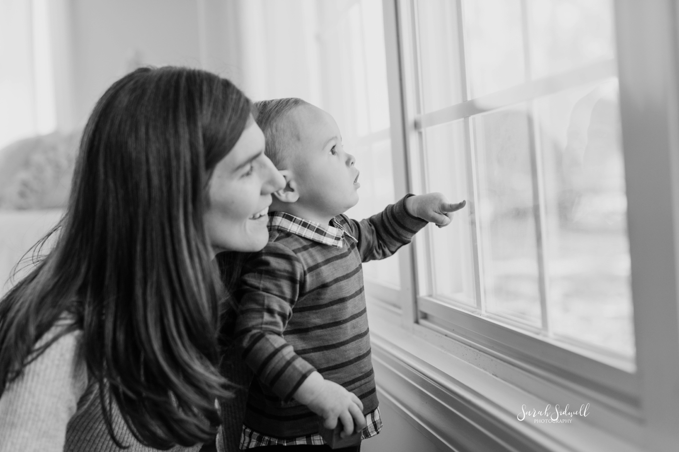 A mother stands behind her toddler, looking out of a big window. 
