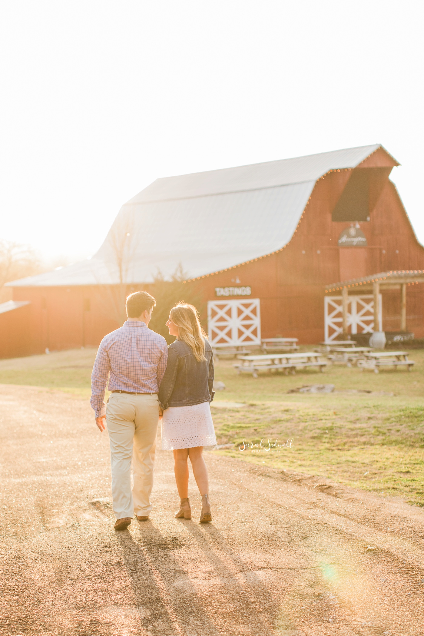 A couple walk in the sunlight towards a barn. 