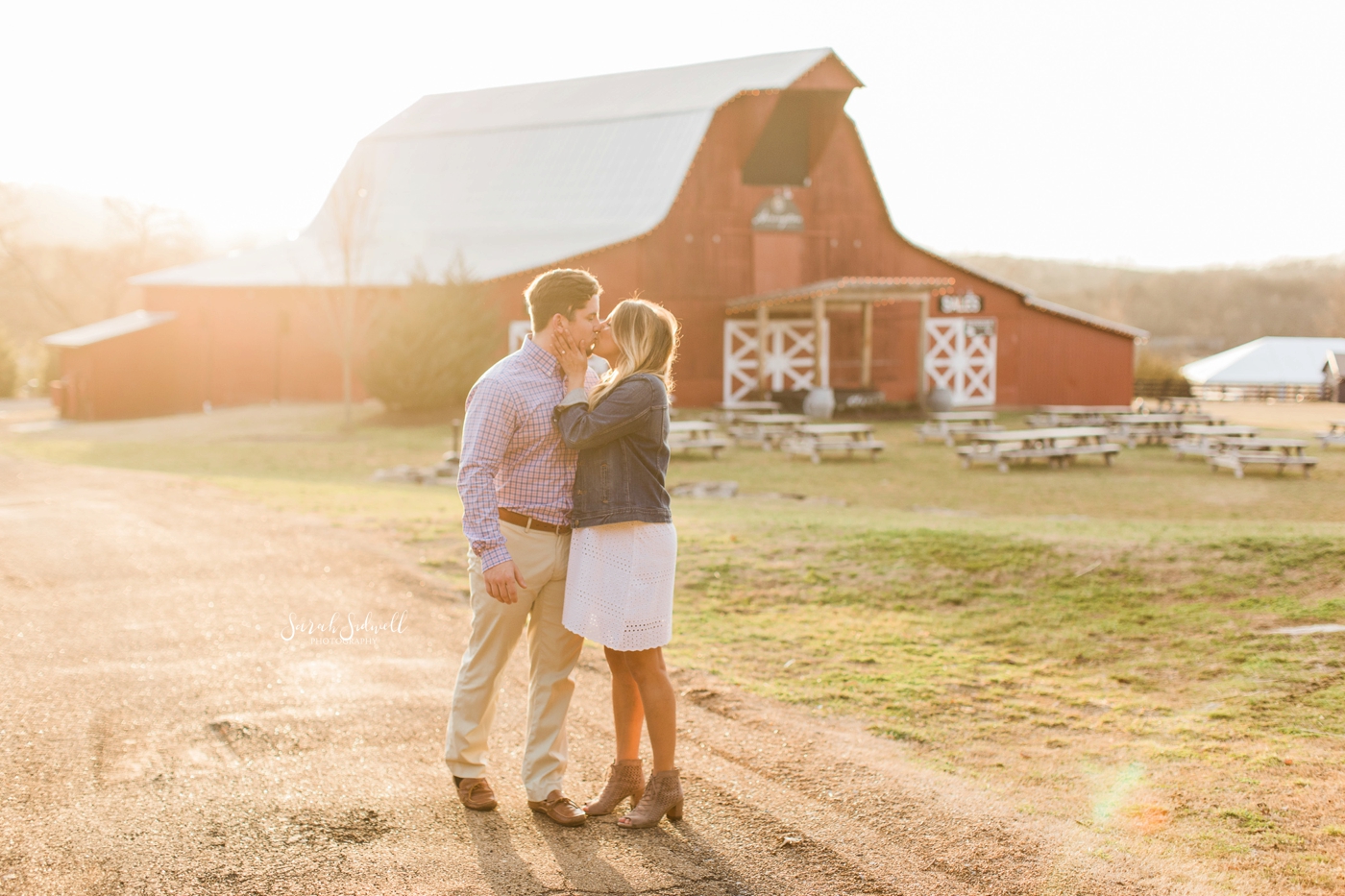 A couple embrace in the sunlight in front of a barn. 