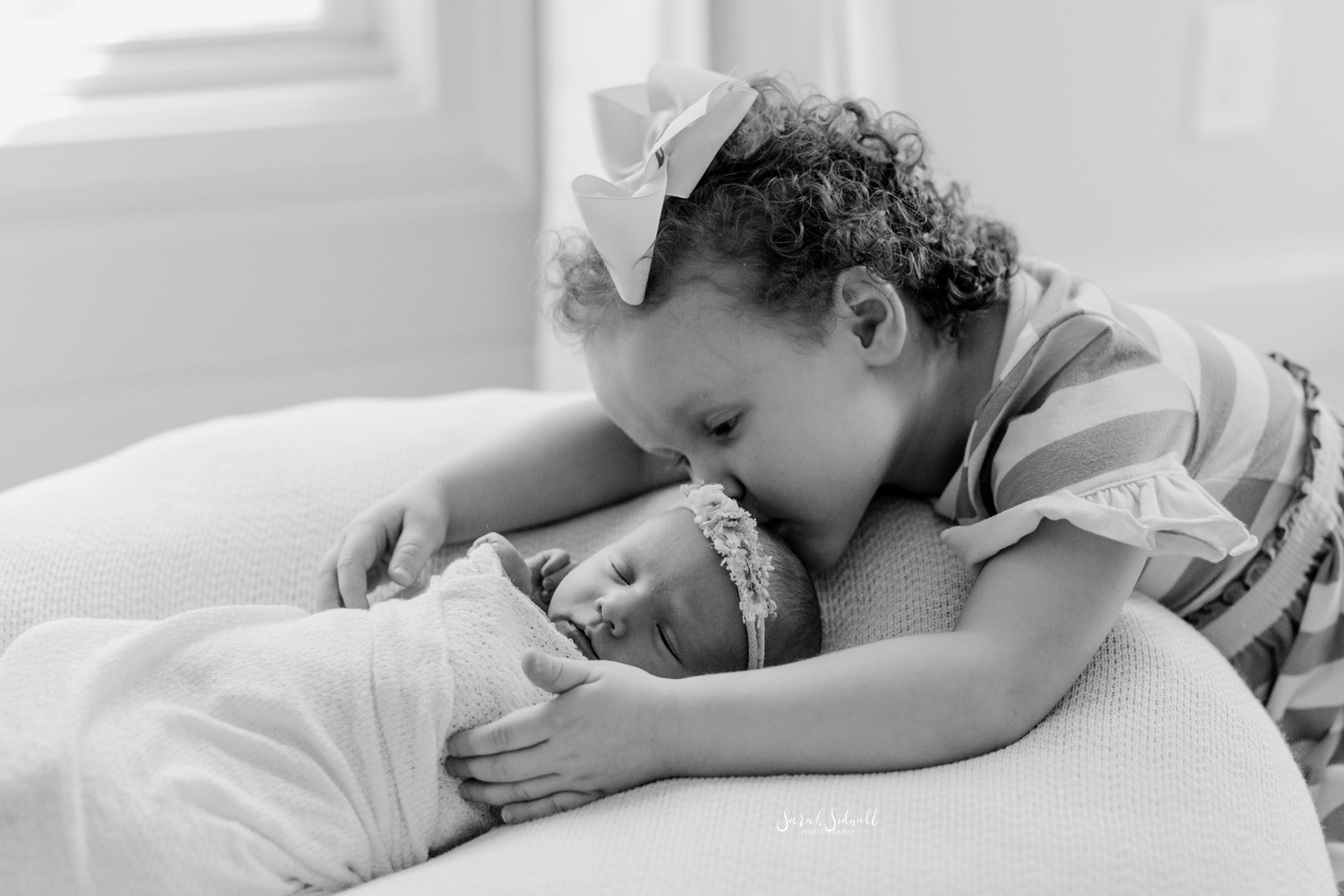 A toddler leans over to kiss her new sister. 