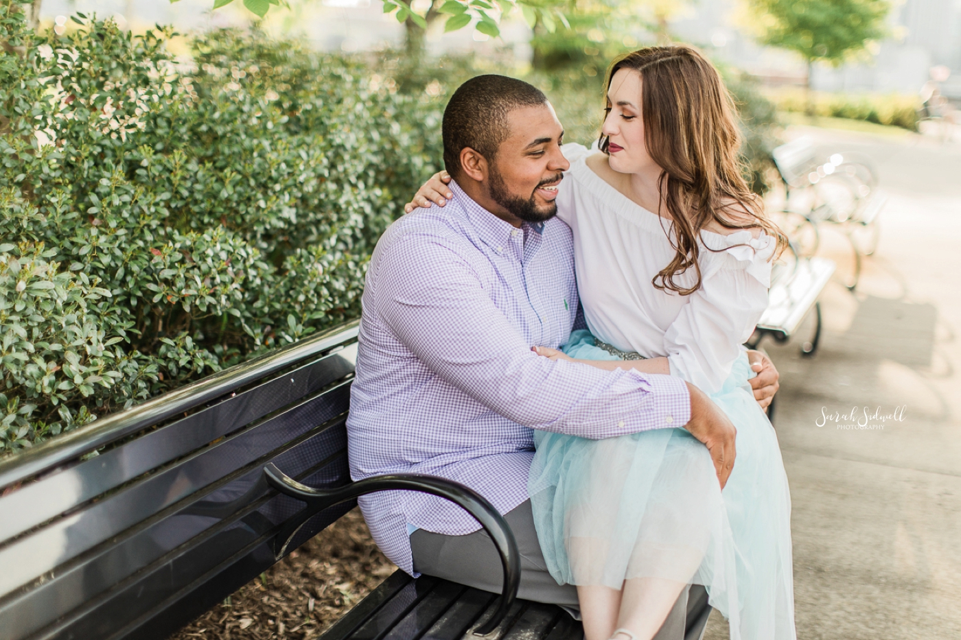 A woman sits on her fiance's lap on a park bench. 