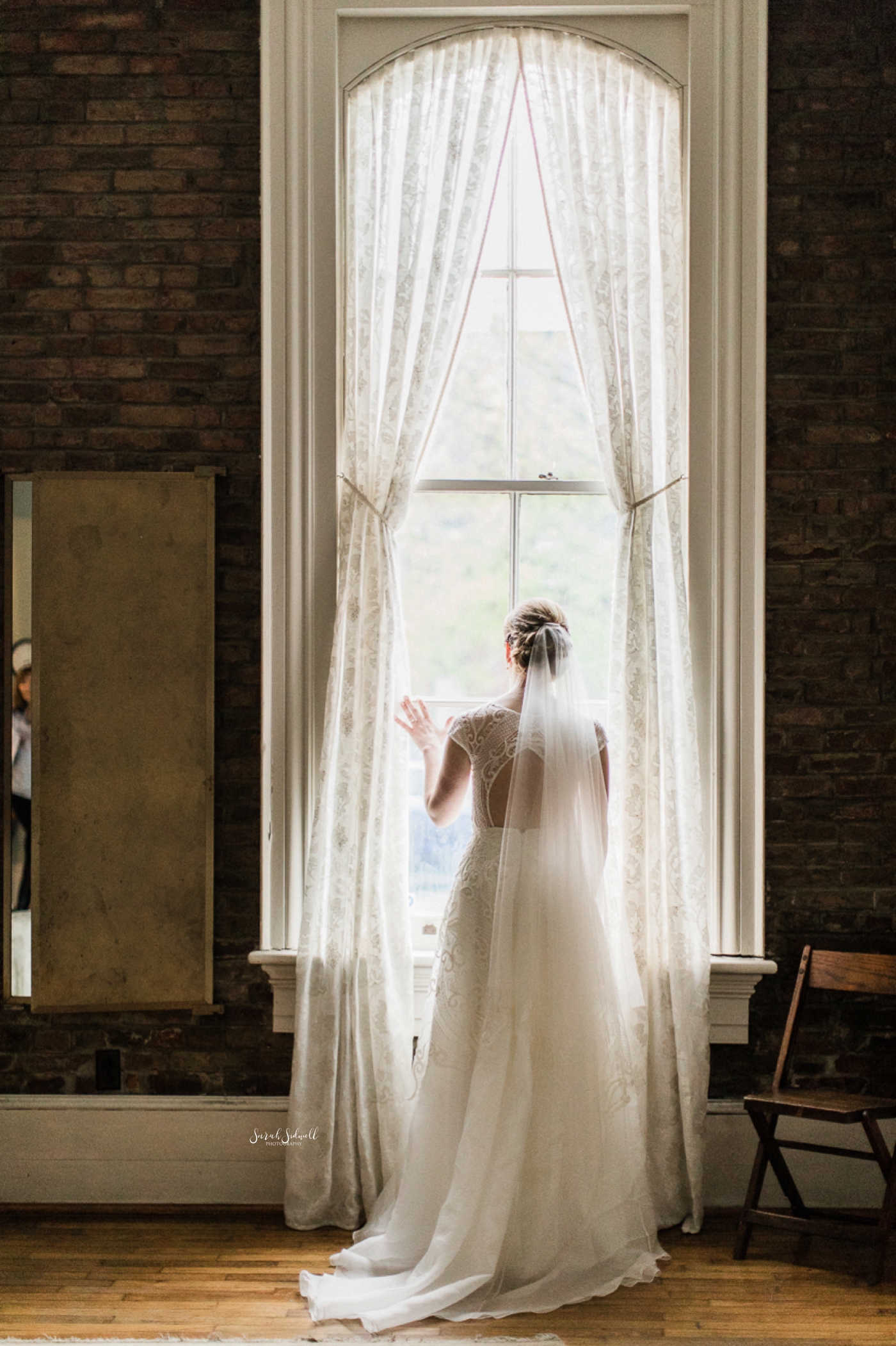 A bride in a white dress stands looking out of a tall window. 