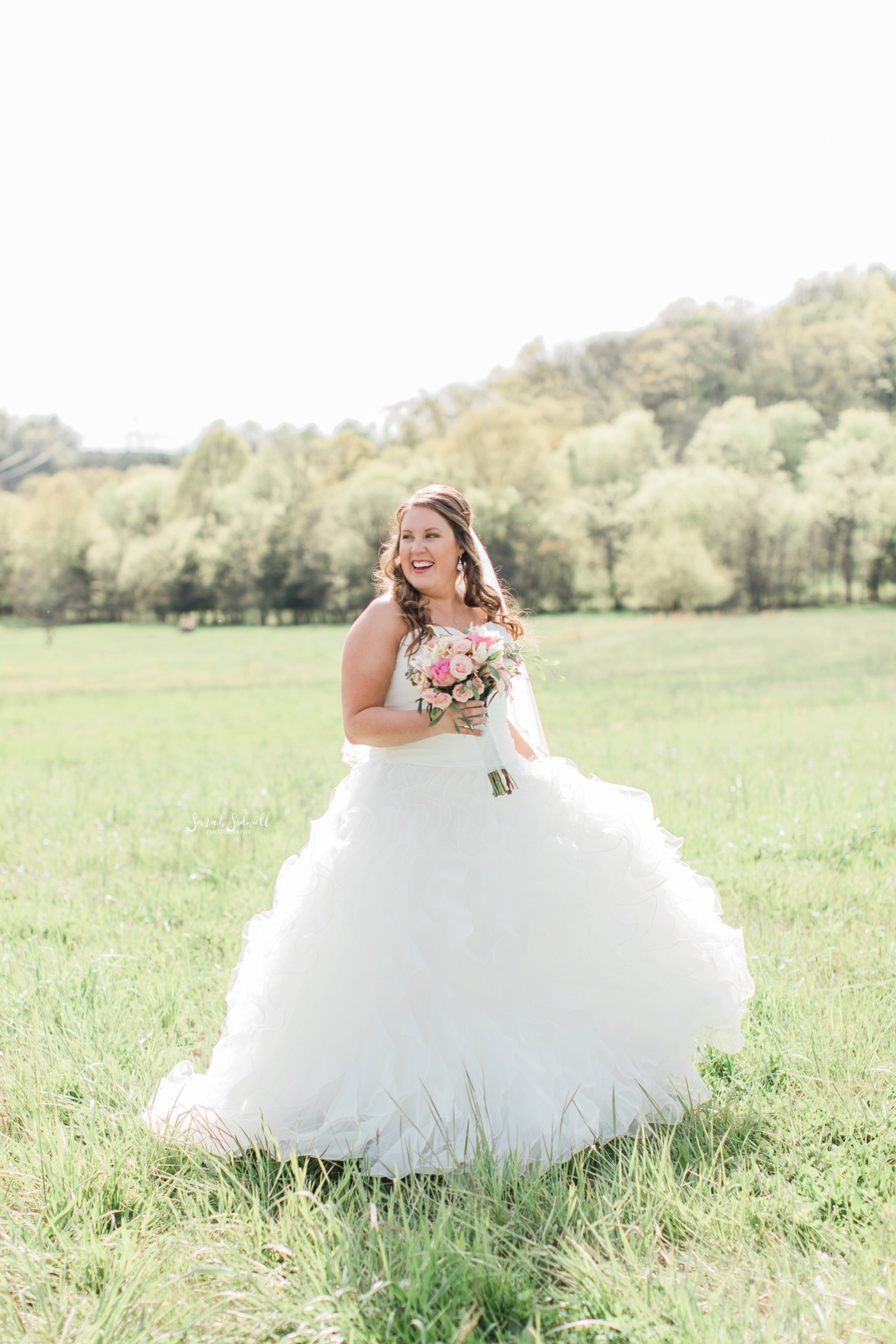 A bride stays in a green field, holding her bouquet. 