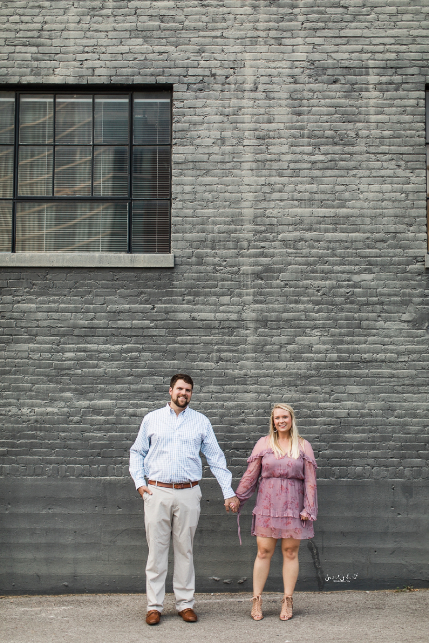 A couple hold hands in front of a grey brick building. 