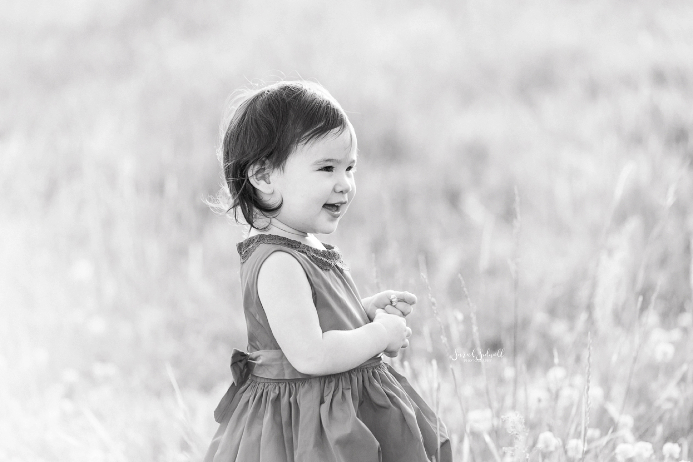 A baby stands in a grassy field, smiling into the distance. 