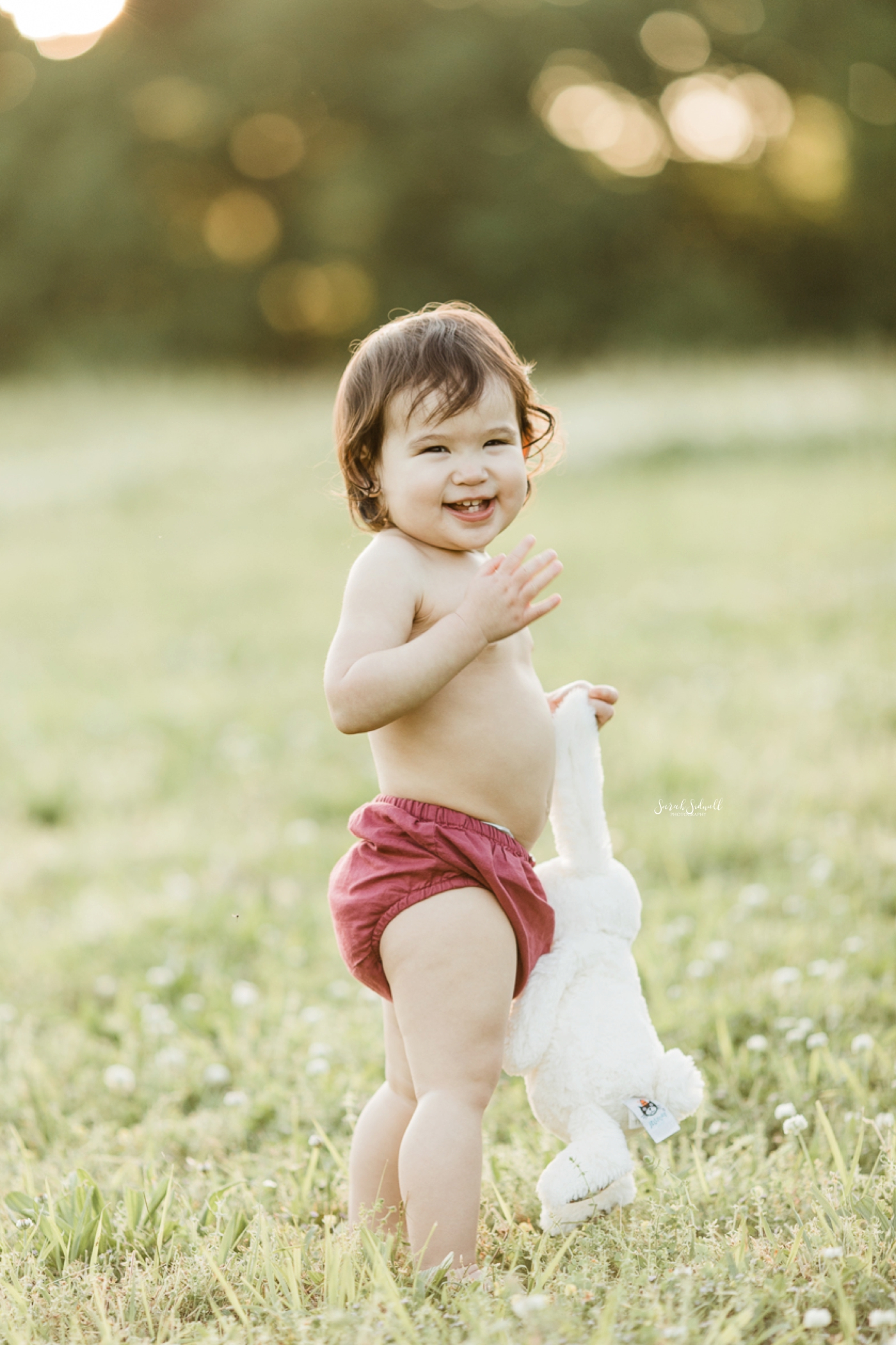 A baby wearing red bloomers holds her dress in her hands. 