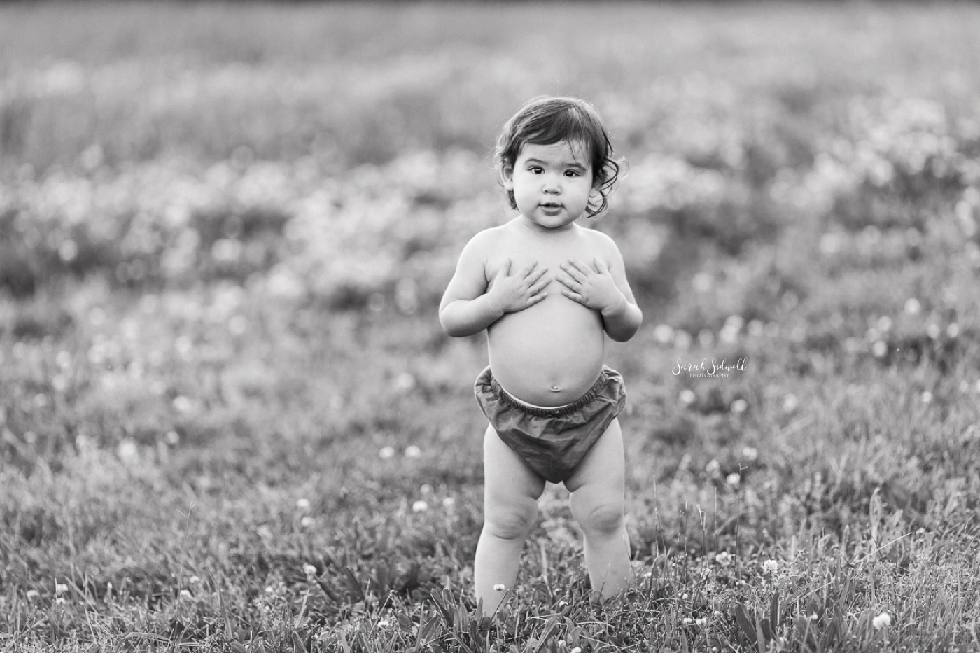 A toddler with a big belly, wearing only bloomers walks outside. 