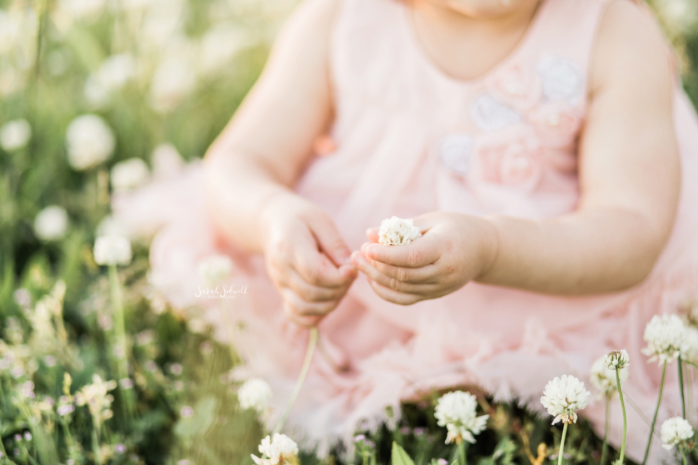A toddler wearing a pink dress bends down to pick some white flowers. 