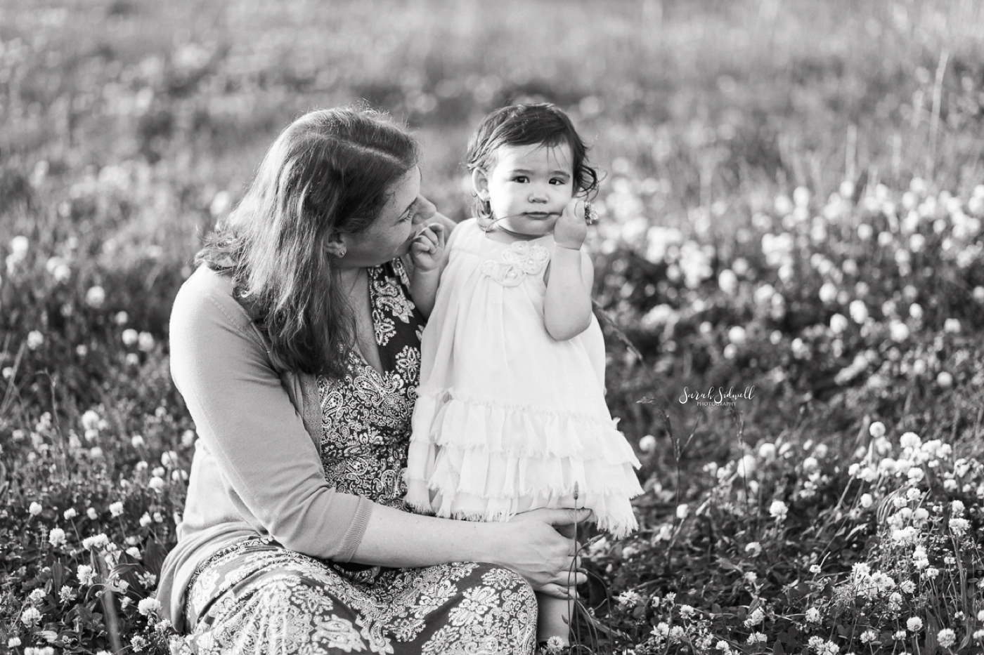 A mother kneels to her toddler as they sit in a field of white flowers. 