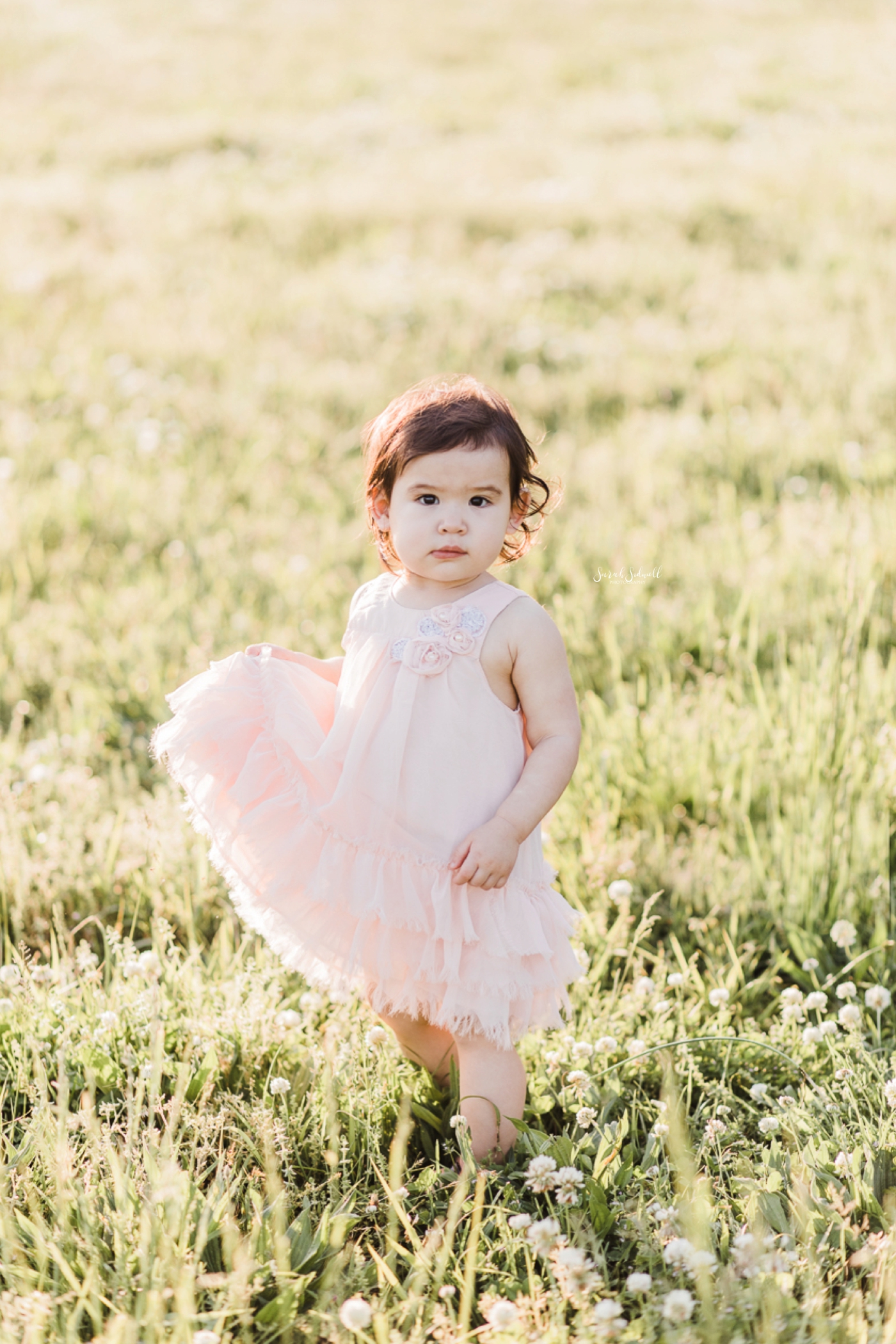 A toddler holds her pink dress in her hands as she walks. 