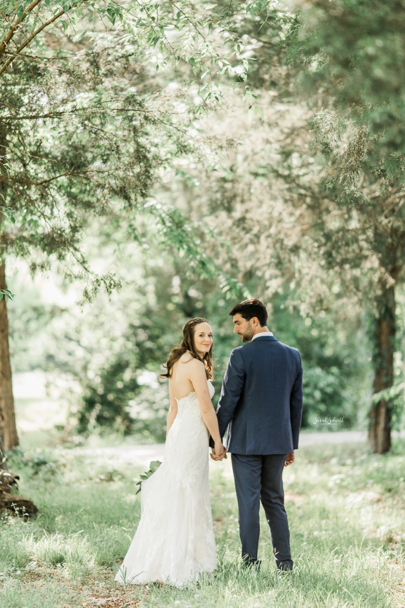 A newlywed couple take a walk under large trees. 