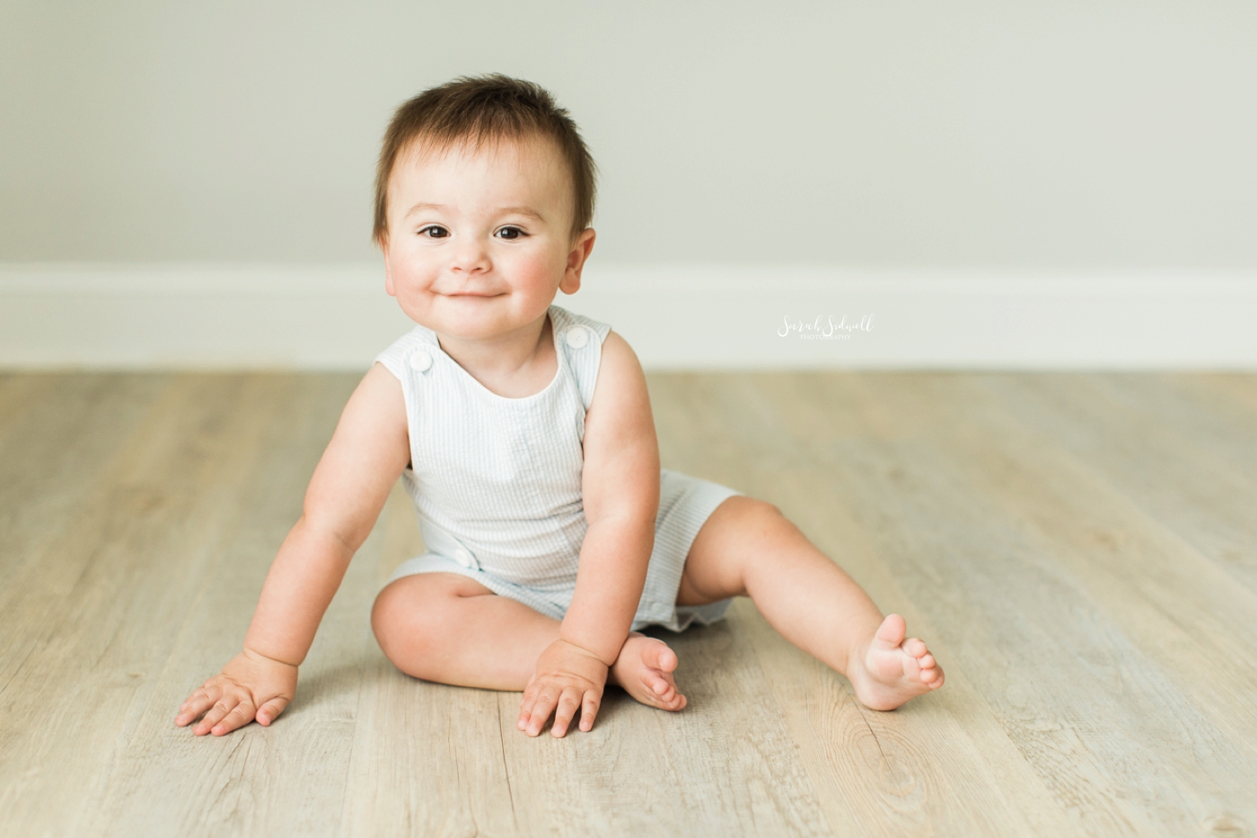 A baby boy sits on the floor and smiles. 