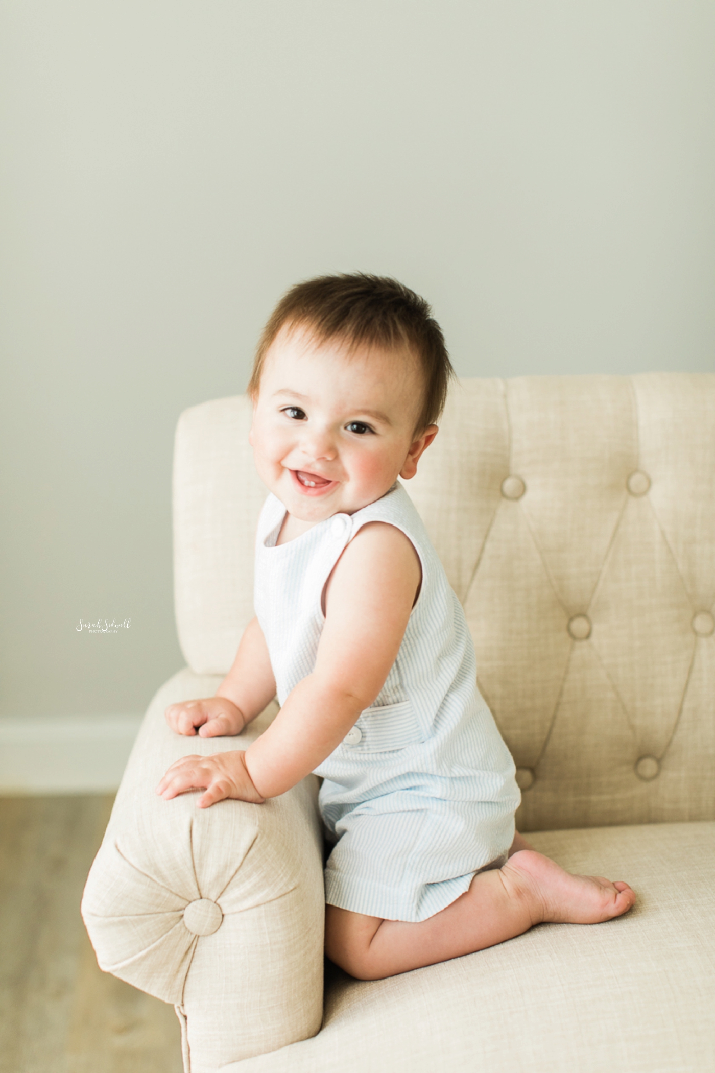 A baby boy sits on a white chair during family photography session. 