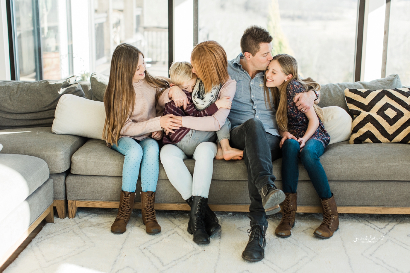 A family of five sits on a couch together and hug each other. 