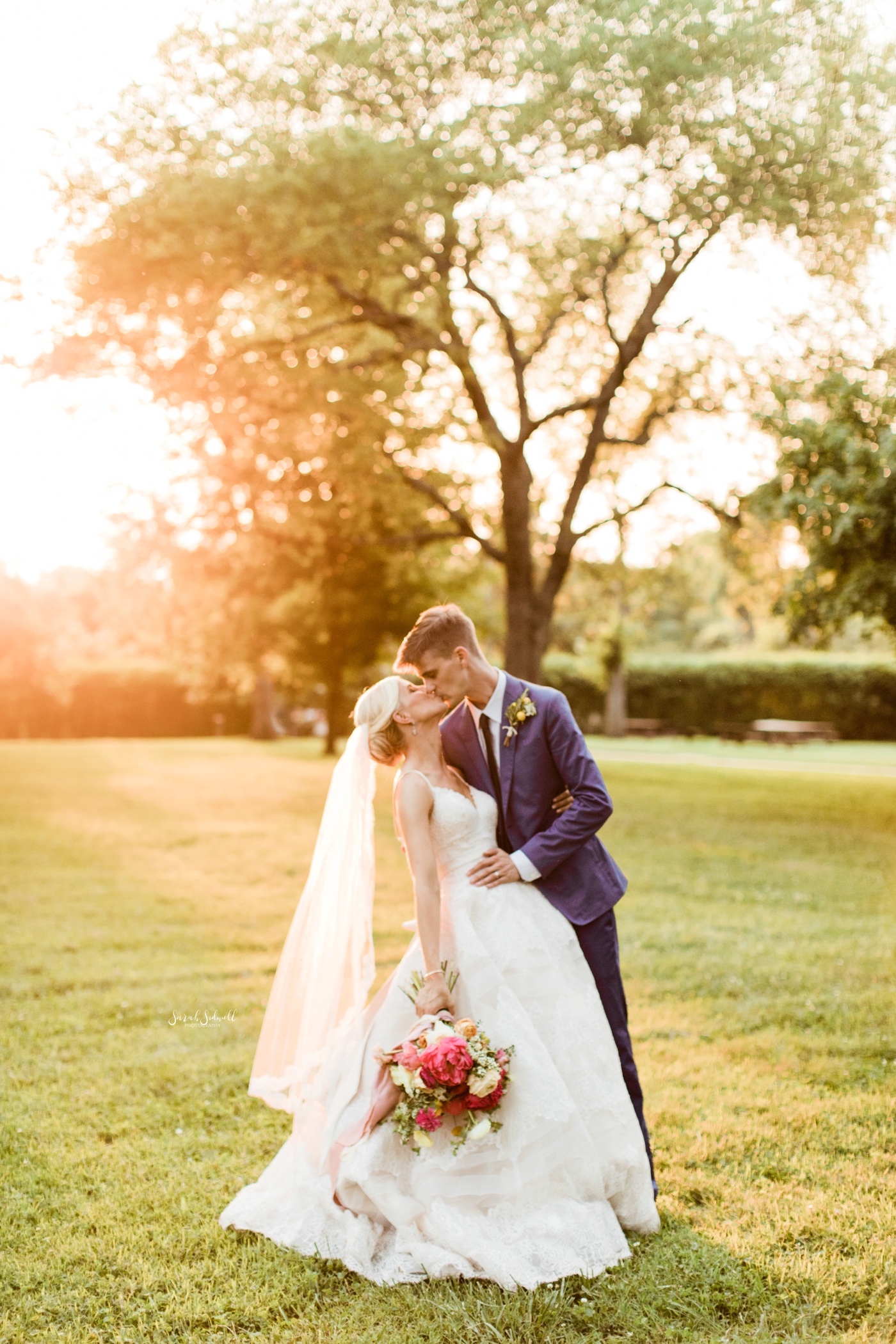 A man kisses his bride in front of a sunset. 