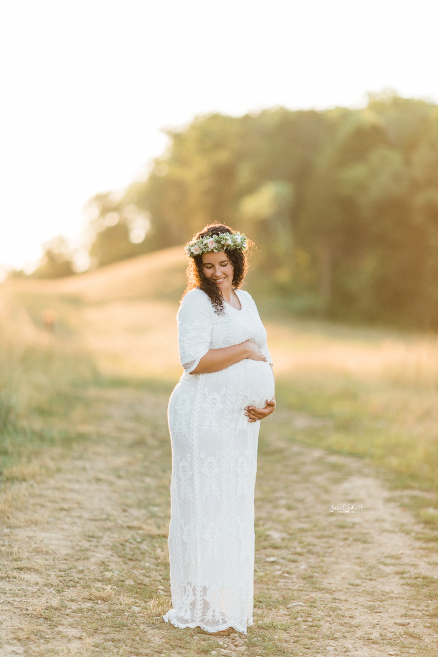 A pregnant woman wearing white and flower crown holds her belly. 