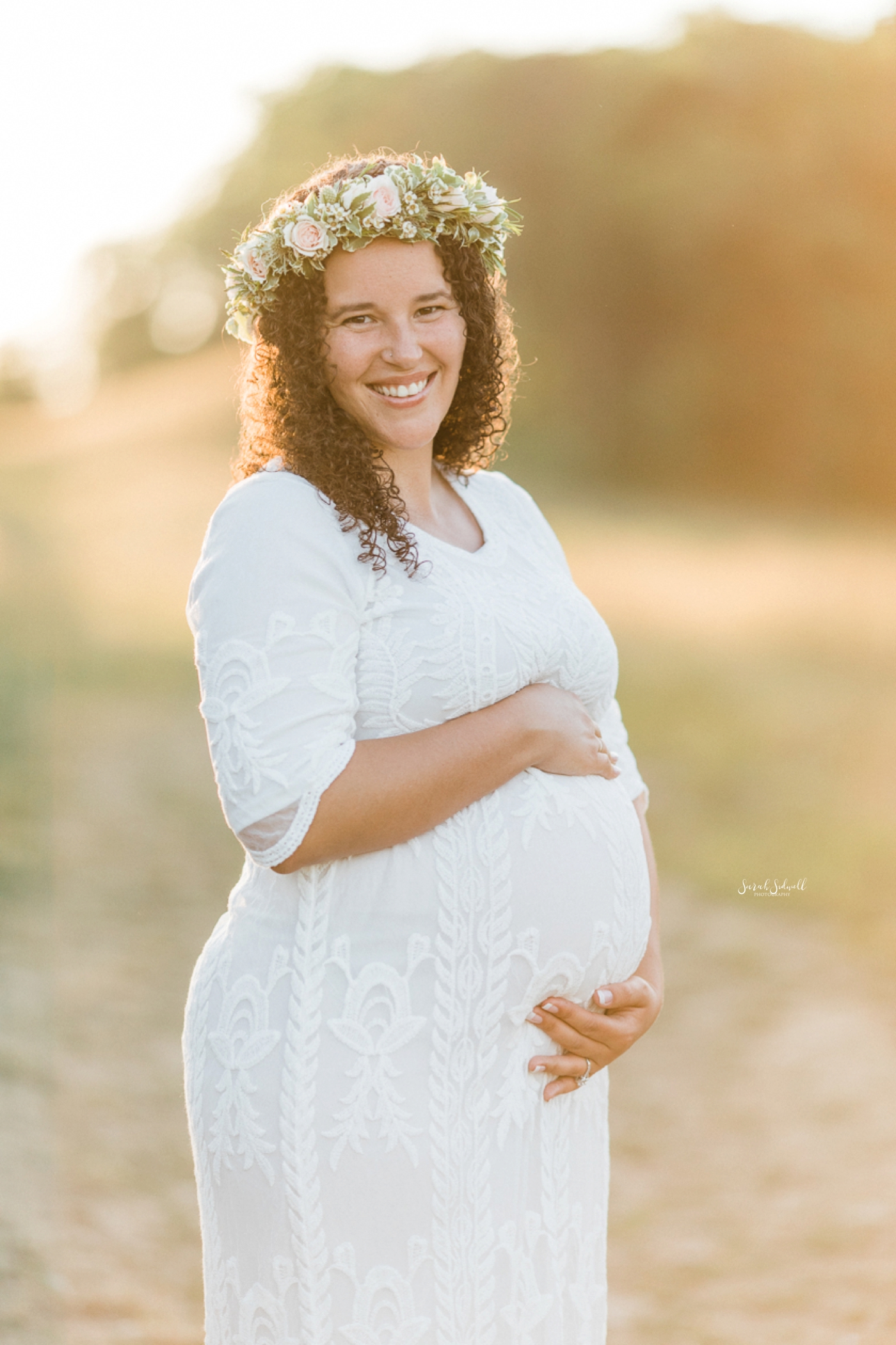 The sunset illuminates a pregnant woman wearing a white dress and flower crown. 