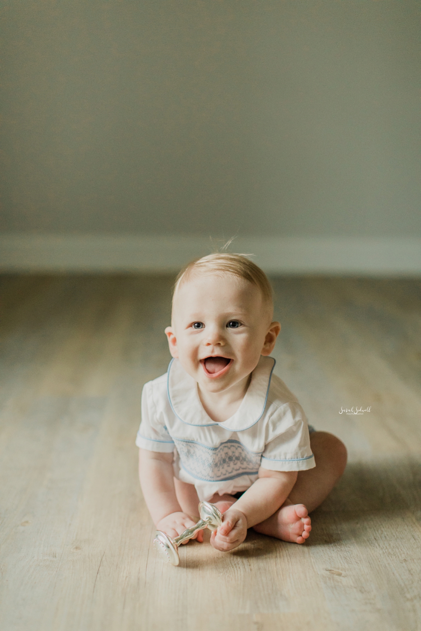 A baby sits up on a wooden floor and smiles. 