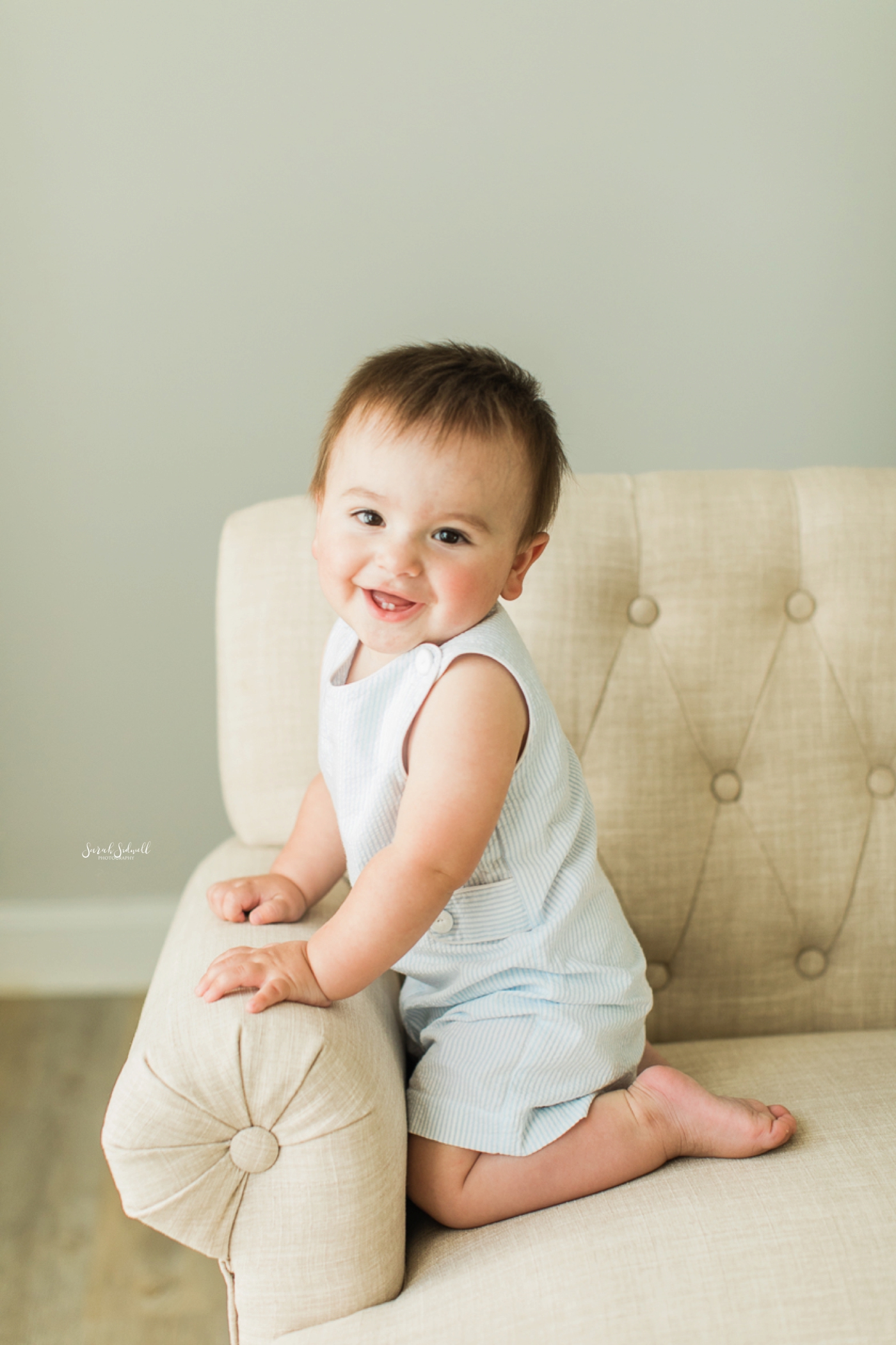 A baby sits on his knees in a white chair. 