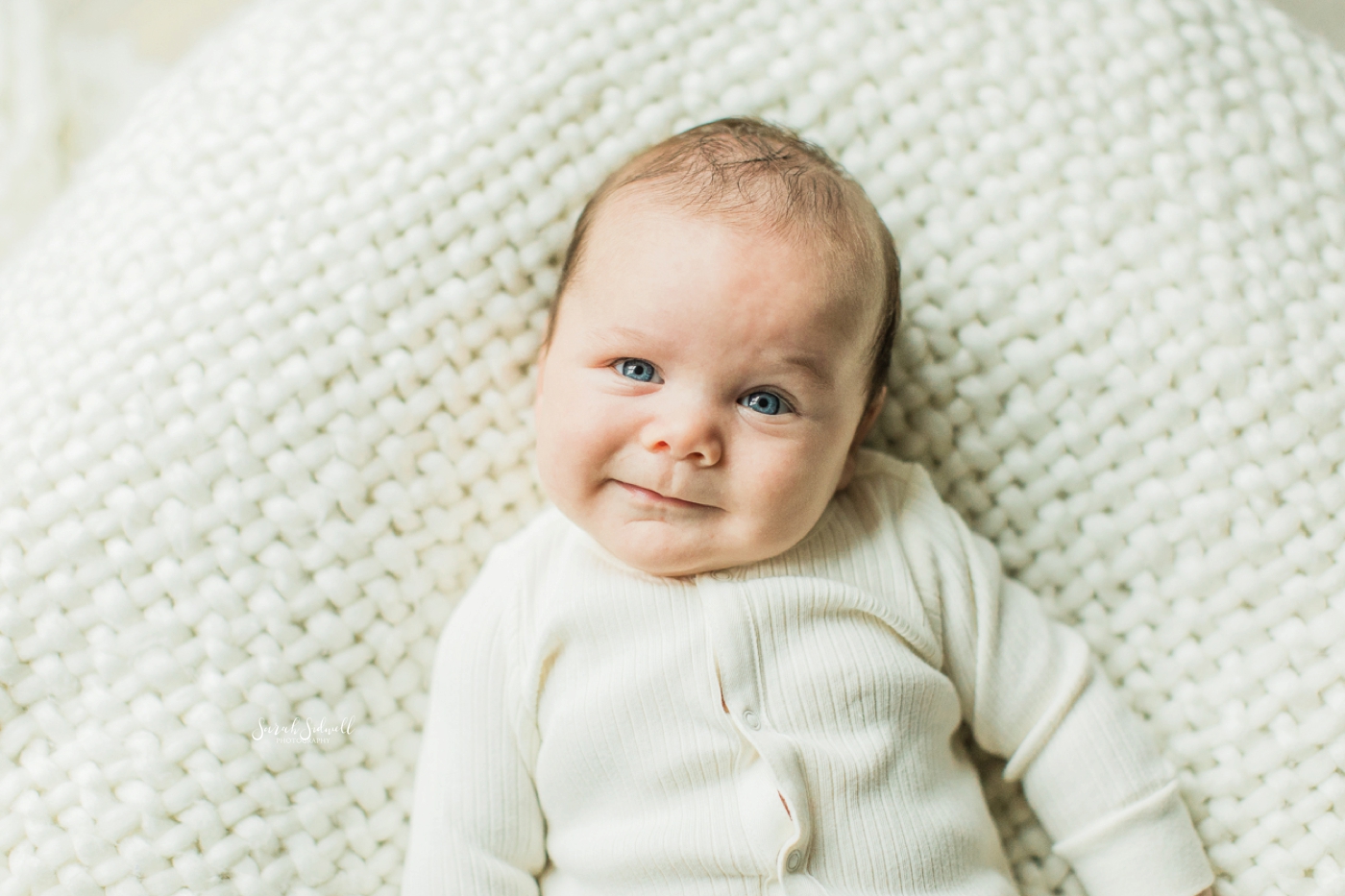 A baby makes a funny face while laying on a white blanket. 