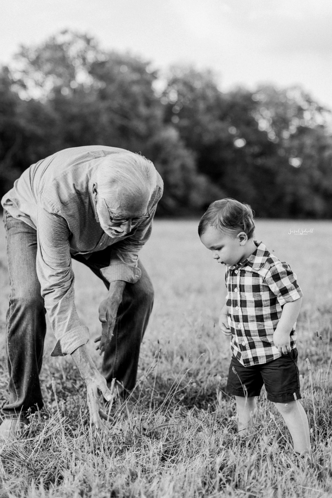 A grandpa bends down to show his grandbaby something in the grass. 