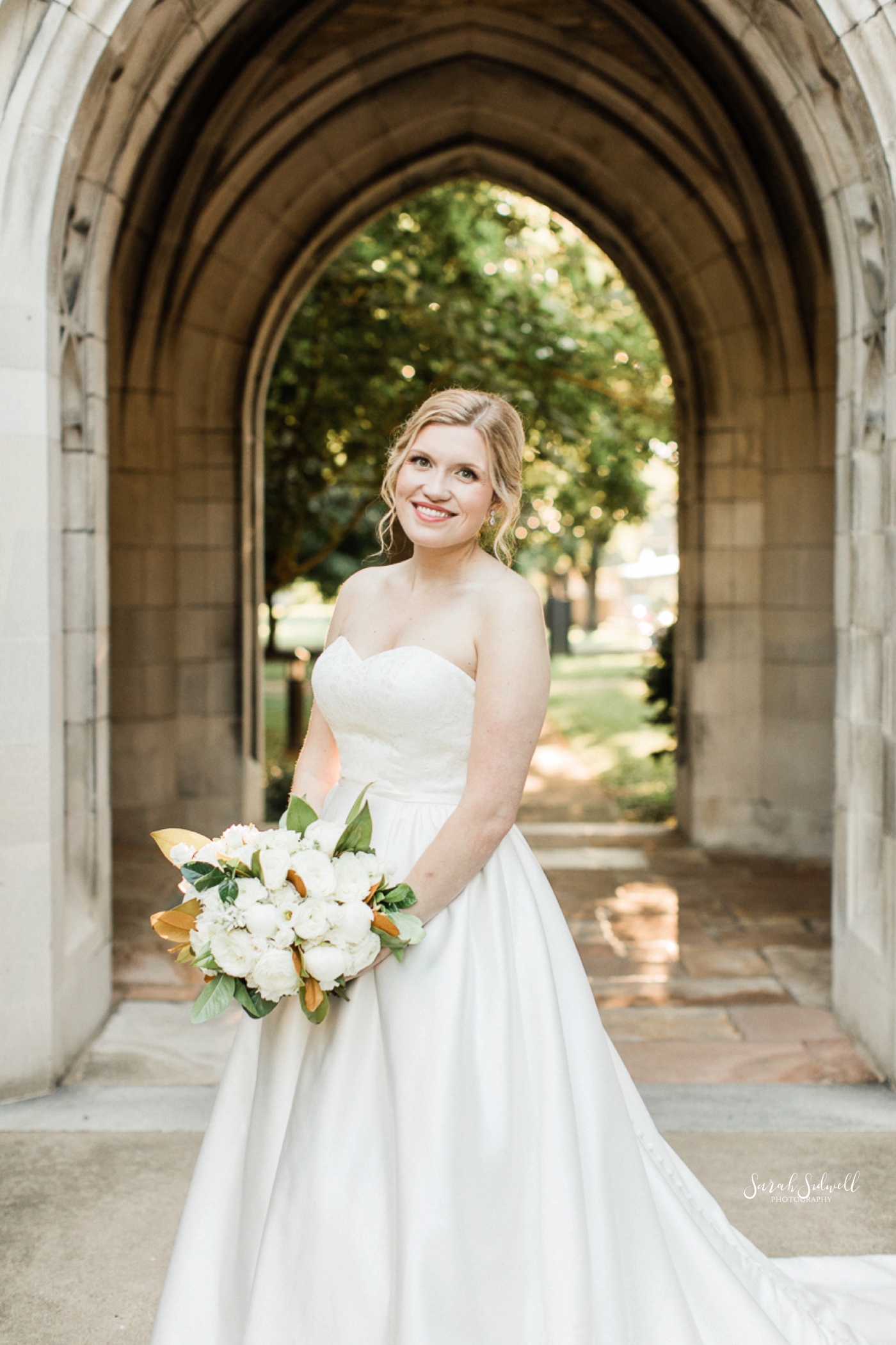 A bride stands under an arch holding her flowers. 