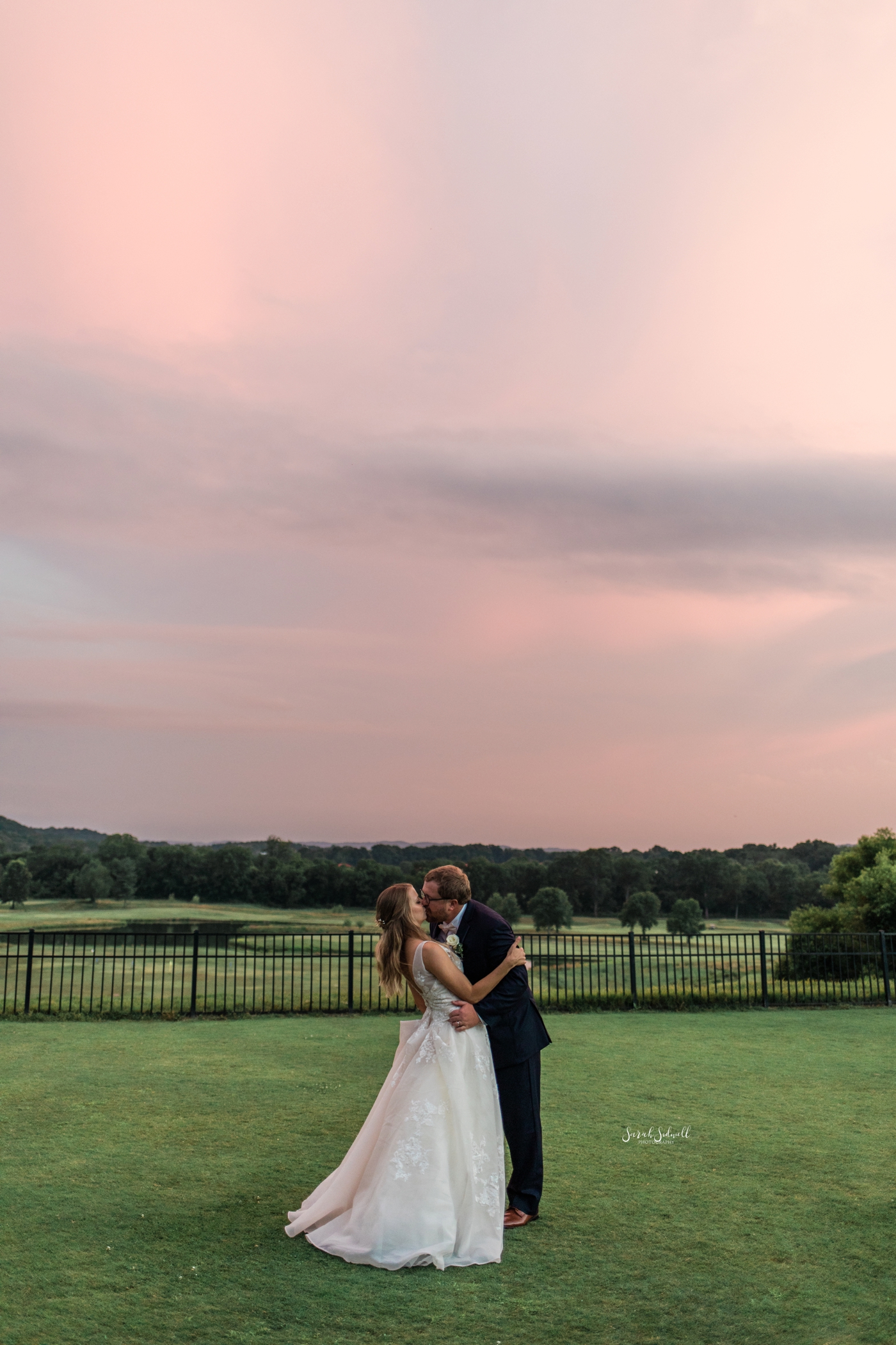 A bride and groom dance in front of a pink sunset. 