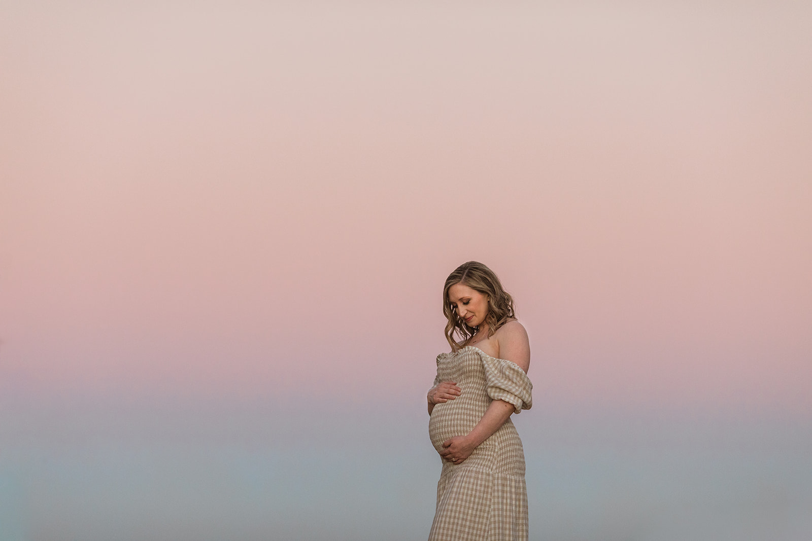 Photo of expecting mama holding her pregnant belly with both hands. Mom wearing a yellow/cream and white checkered off-the-shoulder maxi dress. Pastel pink and blue sky in the background.
