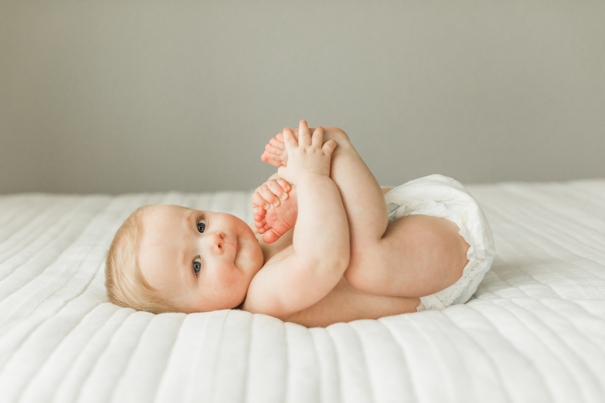 Photo of baby boy laying on his back on bed as he pulls his little legs up to his face and looks at camera. Photographed by Nashville baby photographer in her studio.