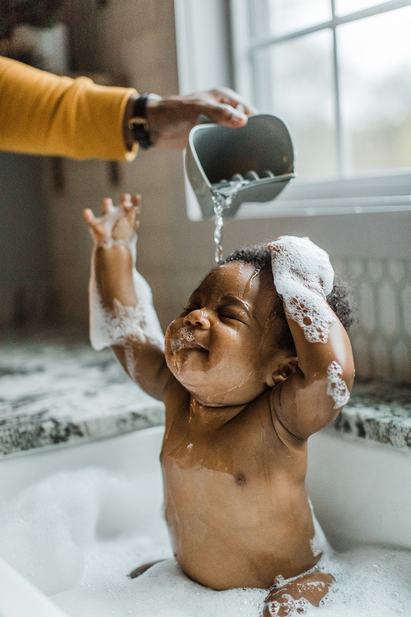 Photo of black baby in kitchen sink filled with water and bubbles. Photo of dad's hand pouring water onto son's head as they wash him off. Baby with eyes closed as waters pours on his head.