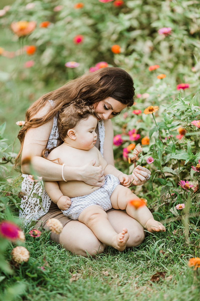 Outdoor photo of baby boy with baby rolls sitting in mamas thighs. Outdoor photo in flower garden, mom picking pink flowers and showing her curious baby boy during their milestone session in Nashville Tennessee.
