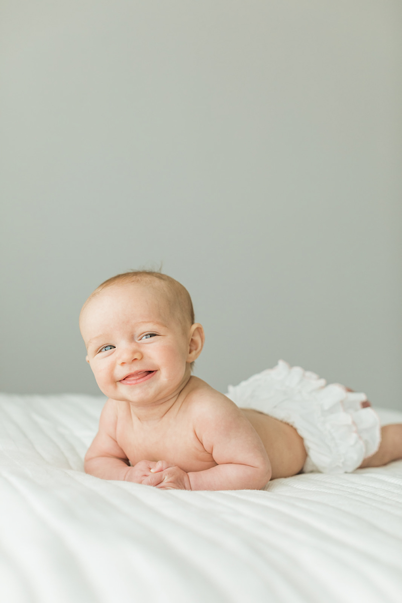 Photo of baby smiling at camera as they are laying down on their belly on bed. Baby milestone photo session photographed by Nashville baby photographer.