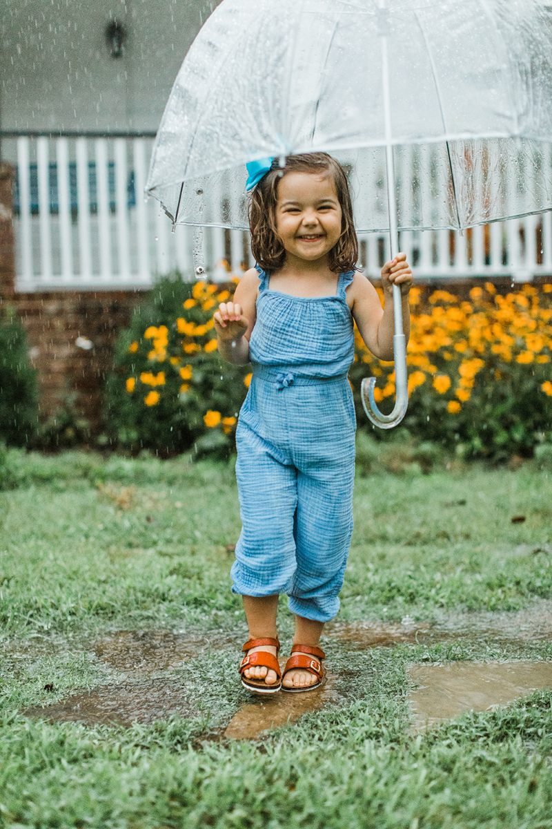 Cute little smiling girl as she hold a clear umbrella while it's pouring outdoors. Little girl wearing blue romper and matching blue hair bow.