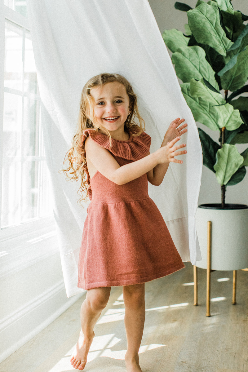 Little girl with natural blonde hair down and wearing a burnt orange dress with ruffle neckline. Young little girl smiling at camera as she plays with white flowy curtains in photo studio. Green tree in white pot in background.