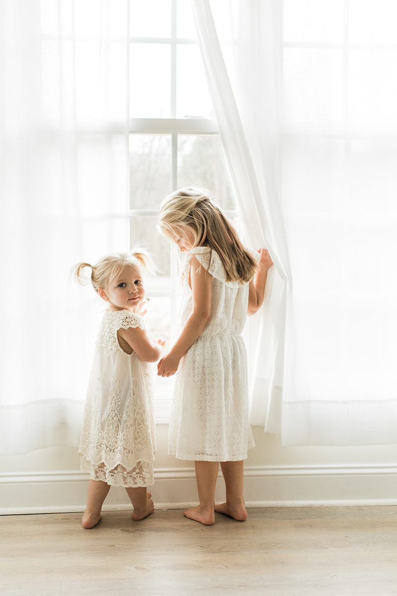 Two young sisters near a white window. Oldest sister holding white curtain and looking down while youngest sister touching the window as she looks at the camera. Both sisters wearing white dresses.