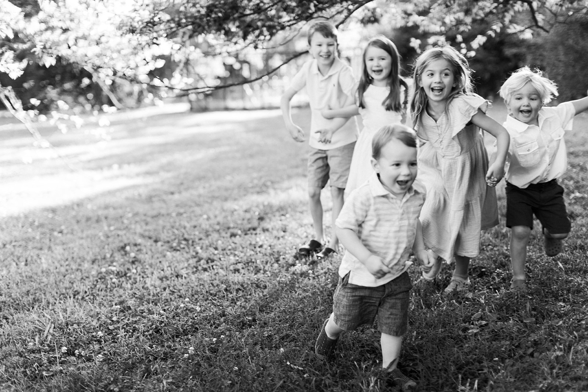Black and white photo of 5 children smiling, laughing and running around outdoors. Two young girls wearing dresses and three young boys wearing shorts and short sleeve shirts.