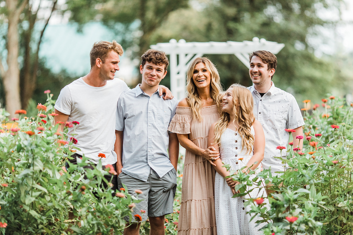 Mom with her four children outdoors in flower garden. Three brothers and young sister. Mom wearing off the shoulder light tan maxi dress and her hair down in loose waves. Young man wearing black shorts and white short sleeve. Two other young men wearing short sleeve button up shirts and grey shorts other young man has tan shorts. Young girl wearing white dress standing in front of mama as she smiles at her.