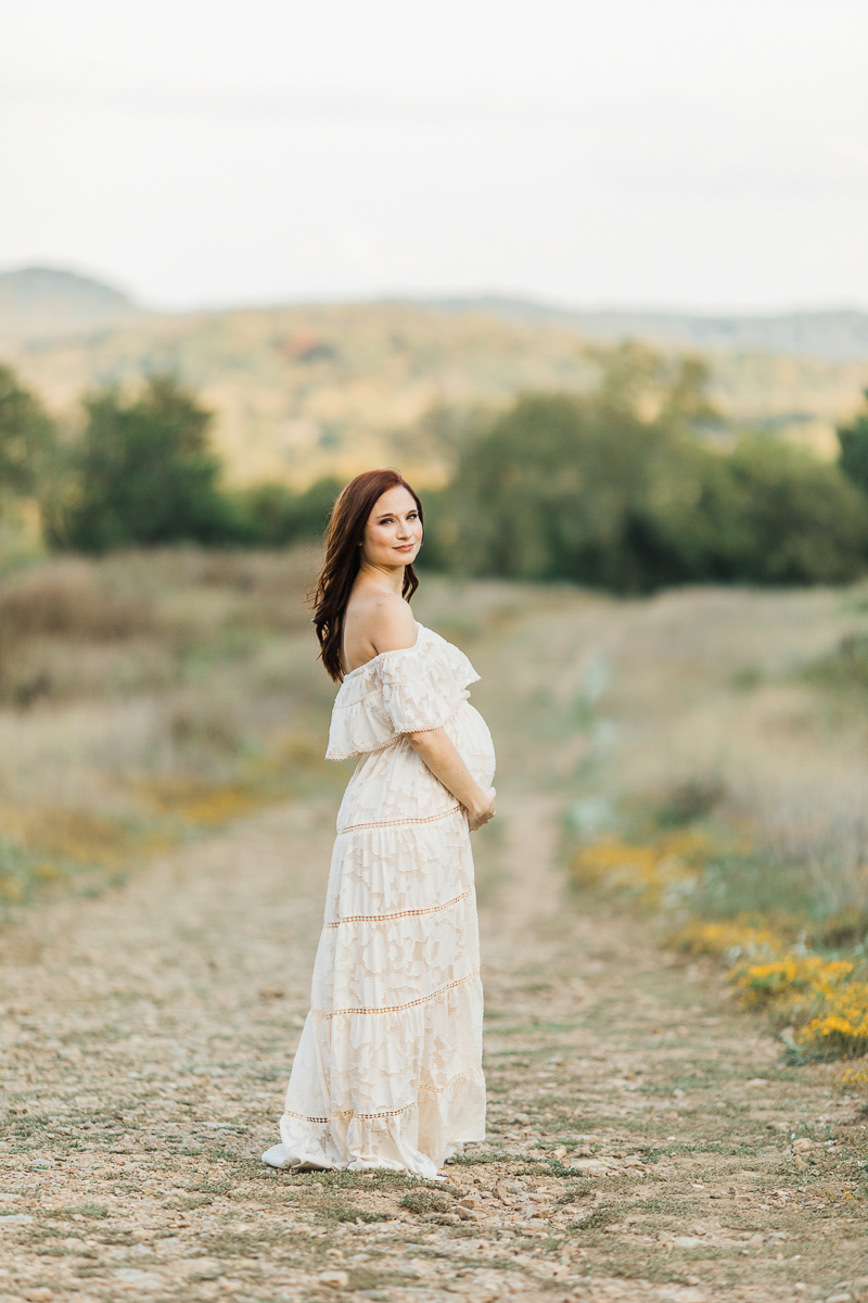 Photo of pregnant woman outdoors wearing a cream colored and laced off the shoulder pregnancy dress. Green outdoors and little yellow flowers on the ground in Nashville Tennessee.