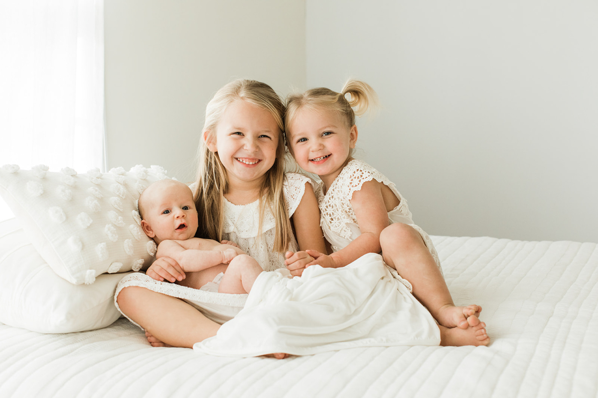 Photo of three sisters. Oldest daughter holding her newborn baby sister as other young sister leans near them. Two big sisters smiling at camera and wearing white dresses. Photographed by Nashville family and newborn photographer..