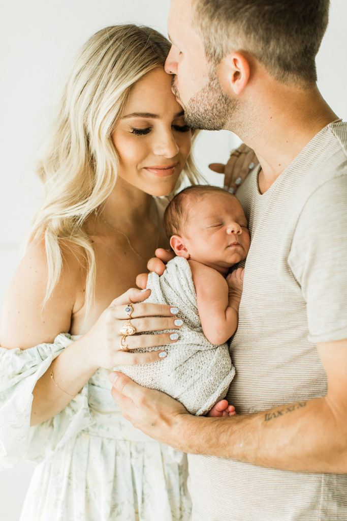 Photo of fashion and lifestyle blogger Hunter Premo and her husband with their newborn baby boy. Husband kissing wife's forhead as he carries and holds their sleepy newborn baby boy near his chest. Mom wearing off the shoulder cream dress  and gold statement rings and dad wearing cream colored short sleeve shirt.