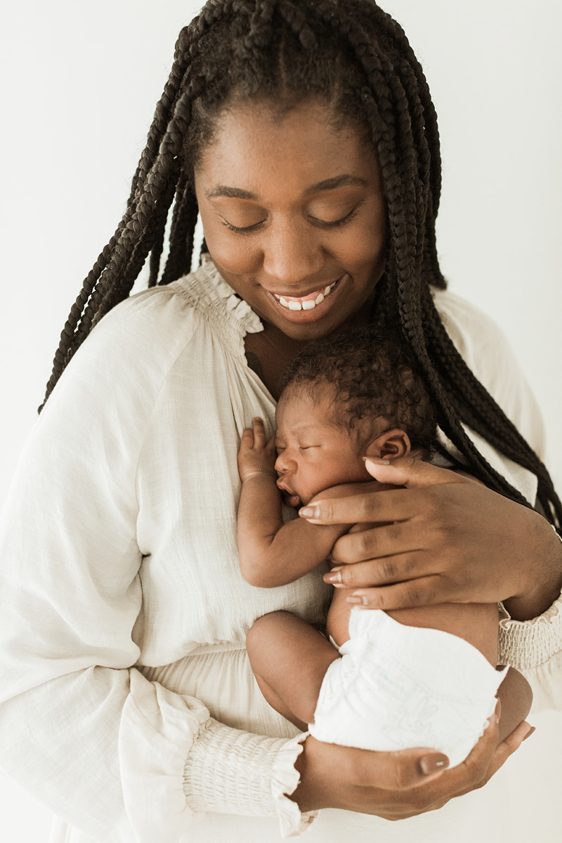 Photo of black mom and little newborn baby boy. Mama holding her newborn baby near her chest as she looks down and smiles at him. Mom wearing cream colored long sleeve dress. Photographed by Nashville newborn photographer, Sarah Sidwell.