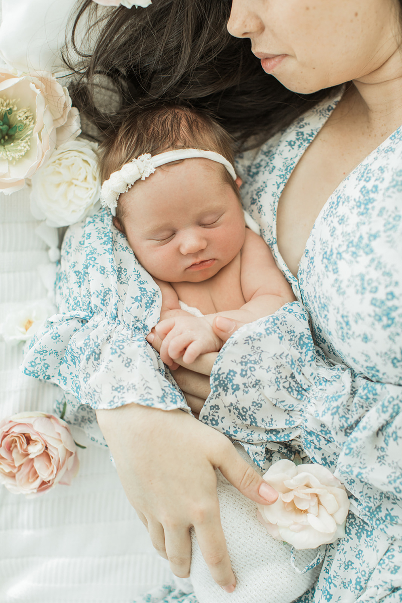 Photo taken from an above angel of mama hugging her sleepy newborn baby girl as they lay on bed. Mama wearing a blue and white floral printed dress. Baby girl wearing little headband with white flowers. Flowers laying around mama and baby on bed.