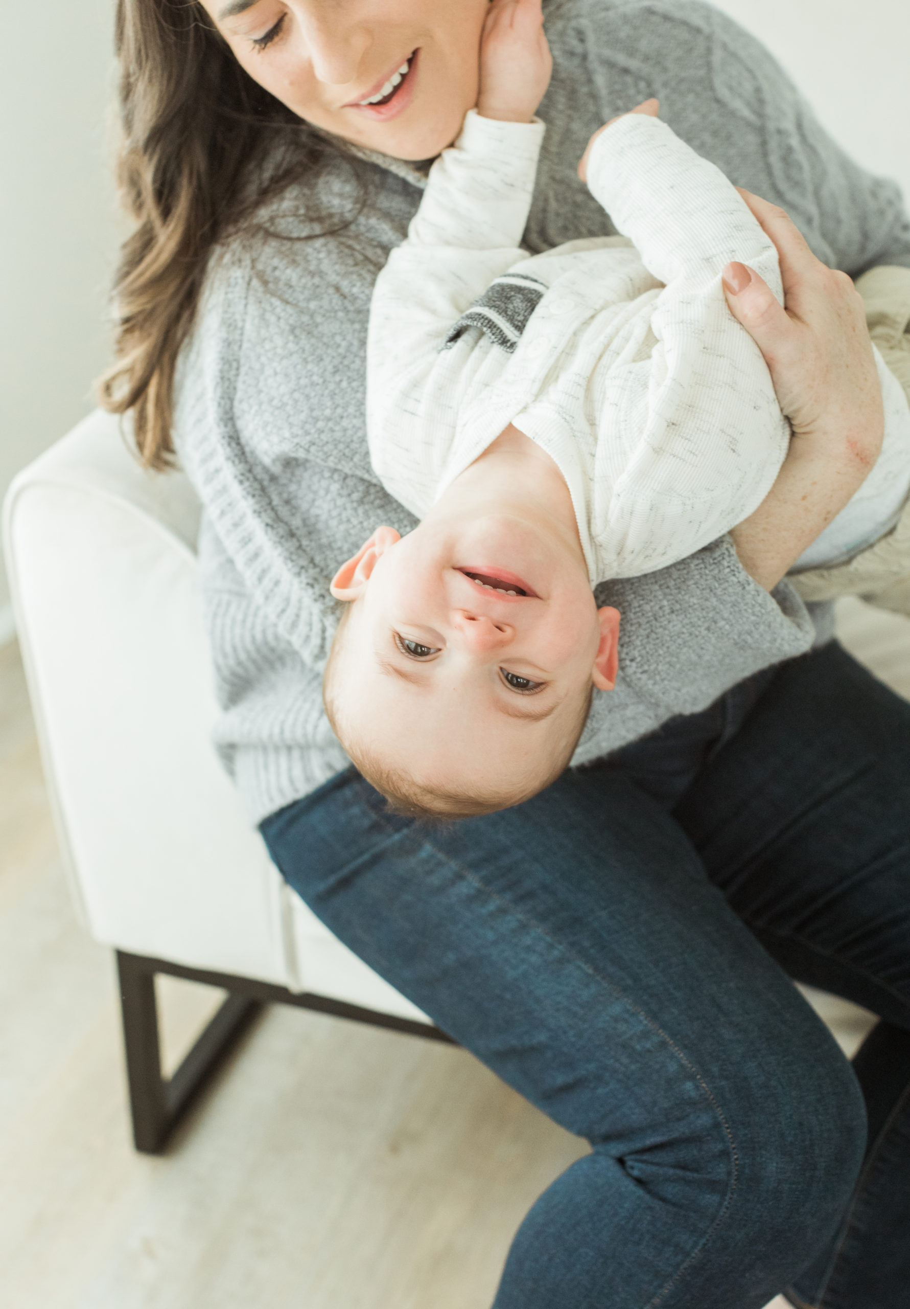 Photo of one year old little boy in mama's arms. Mama sitting down in cream chair, little boy hanging upside down, smiling and looking at camera. Boy wearing light grey long sleeve shirt and khaki pants. Mama wearing grey long sleeve sweater and jeans.
