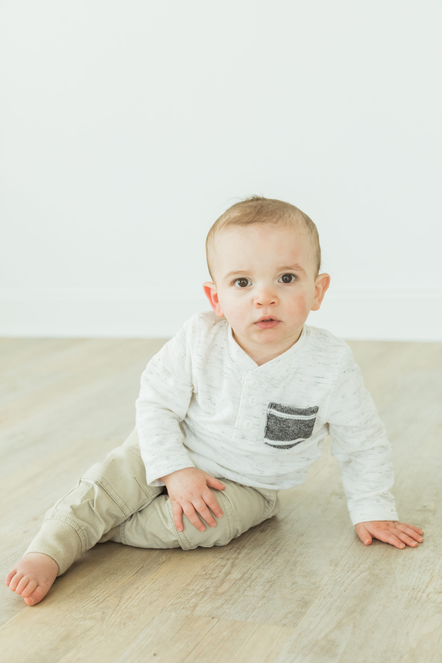One year old little boy wearing long sleeve shirt and khaki pants, barefoot sitting on the light wooden floor.