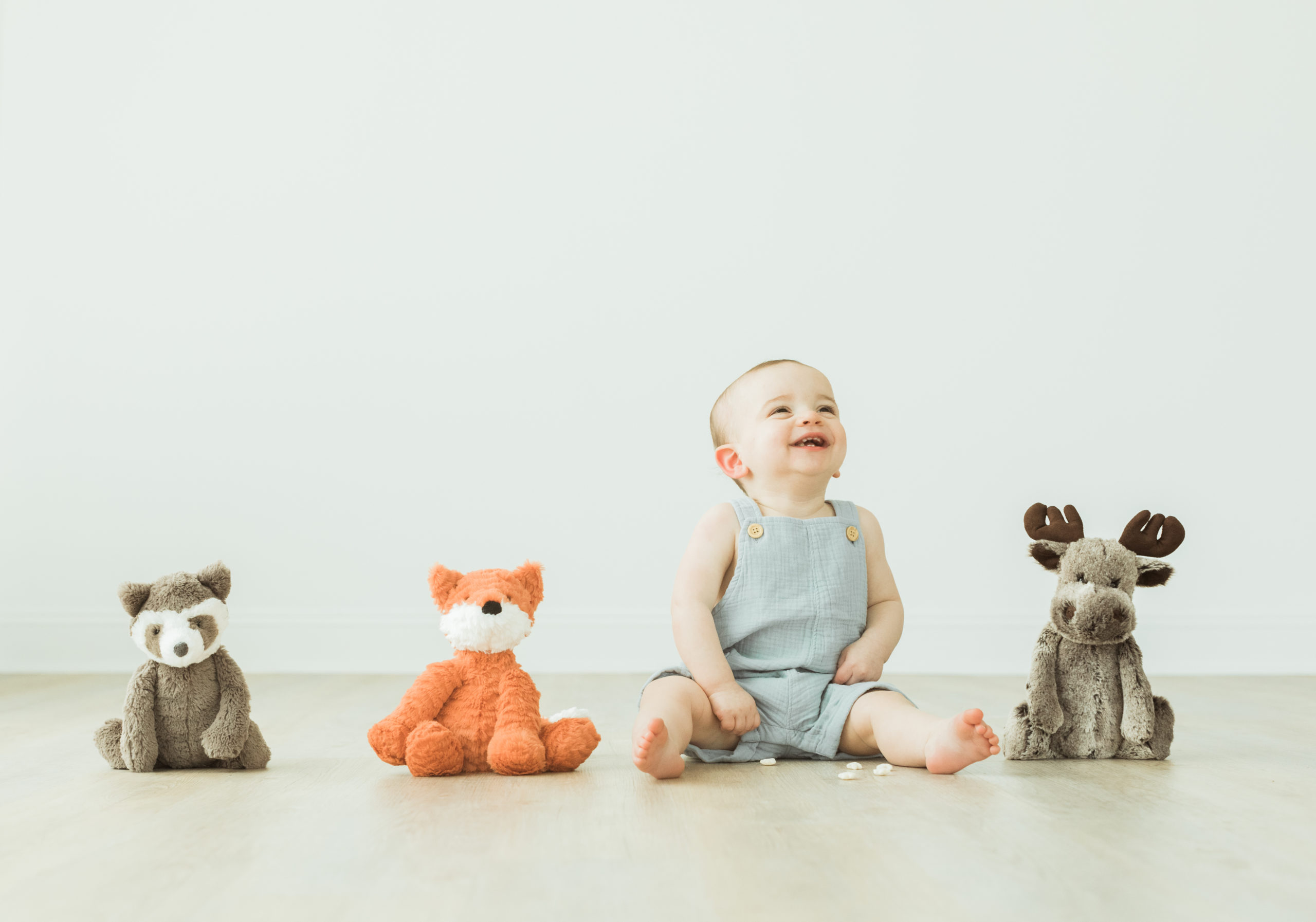 Photo of one year old baby boy sitting down on the floor. Three stuffed animals sitting next to little boy on the floor in a line. Baby boy laughing and wearing a light blue linen overall.
